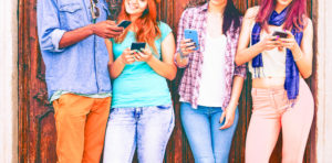 Group of teenagers texting mobile phone messages leaning against urban wall - Row of best student friends using smartphone wearing summer trendy clothing.
