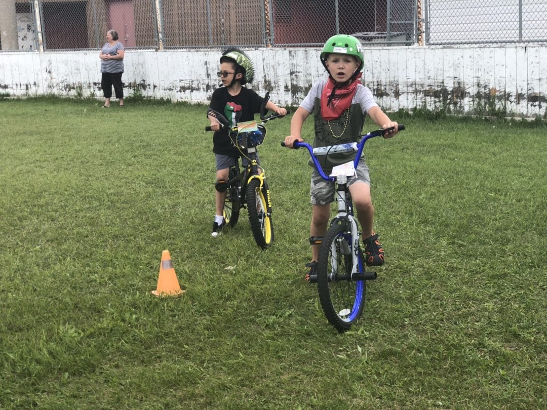 Kids in green helmets riding their bike on grass around a small pylon.