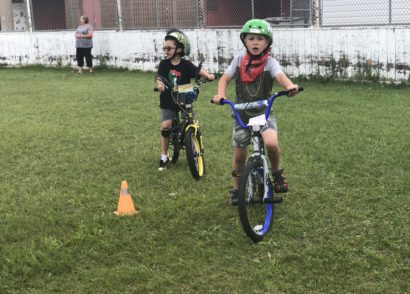 Kids in green helmets riding their bike on grass around a small pylon.
