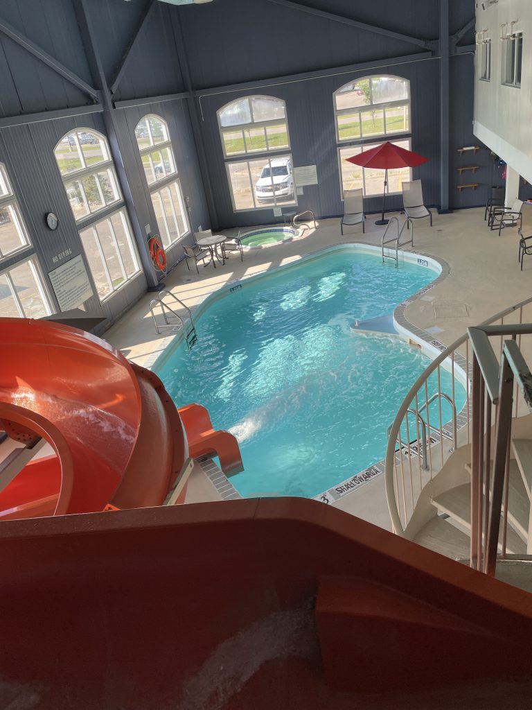 Orange water slide in foreground, feeding into kidney shaped pool and small hot tub at the far side. Orange beach umbrella over deck chairs and orange rescue float match the water slide. Dark blue walls around large, sun-filled windows on the exterior walls. Interior walls are white.