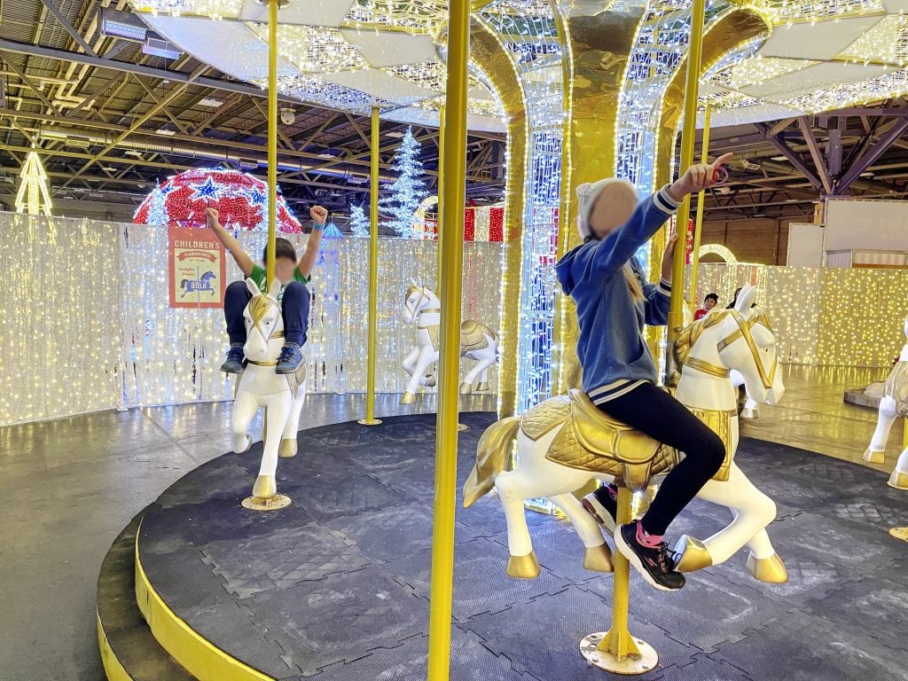 Children riding the white horses on a gold and white light merry go round at Christmas GLOW Edmonton.