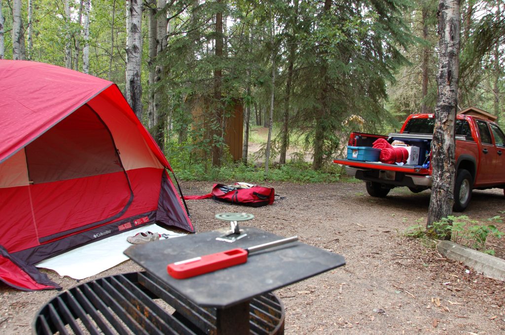 Campsite in the process of being set up with a red tent to the left, its bag on the ground in the middle and an orange truck with a rolled sleeping bag and other supplies in the bed. A fire pit with a red lighter is in the foreground.